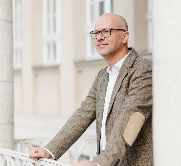 Immobiliengutachter steht auf einem Balkon vor klassischem Gebäude und blickt ruhig in die Ferne.